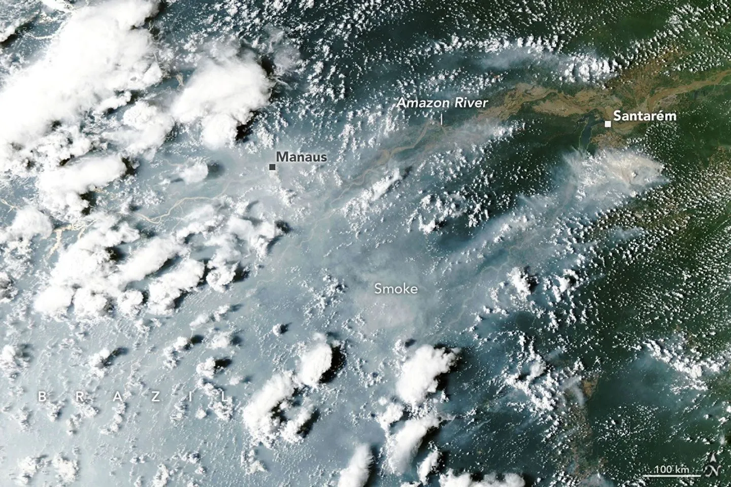 Thick gray smoke partially obscures the Amazon River and surrounding rainforest near the cities of Manaus and Santarém. Puffy white clouds rise above the smoke on the left side of the image.