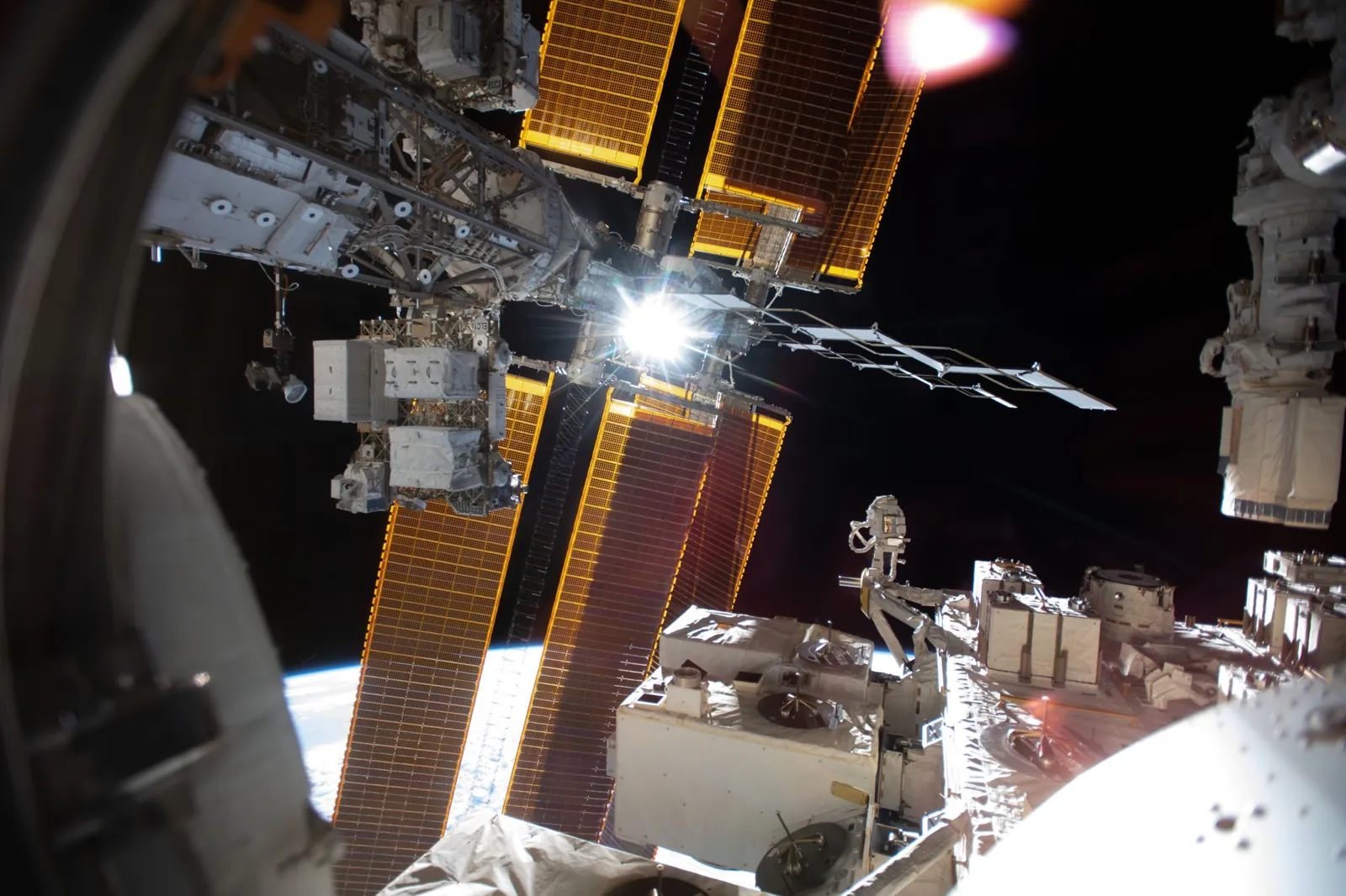 Illuminated solar arrays and structural trusses of the International Space Station are shown against the darkness of space, with sunlight flaring near the center of the frame and Earth’s curved horizo