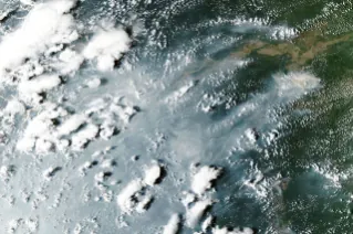 Thick gray smoke partially obscures the Amazon River and surrounding rainforest near the cities of Manaus and Santarém. Puffy white clouds rise above the smoke on the left side of the image.