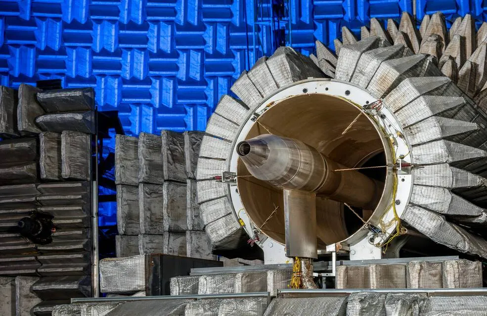 A Learjet engine nozzle peeks out from NASA Glenns Nozzle Acoustic Test Rig, a cylinder surrounded by tan wedges. The walls of the Aero-Acoustic Propulsion Facility, also covered in wedges, glow blue 