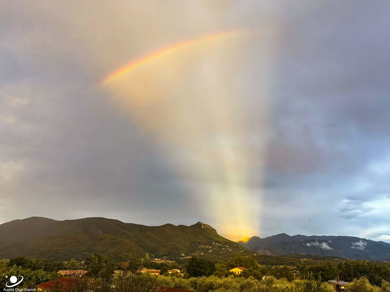 Hills are seen in the distance below a partly cloudy sky. A group of orange rays appears to eminate from one area behind the hills. Across this fan of rays, near the top of the image, a short rainbow 