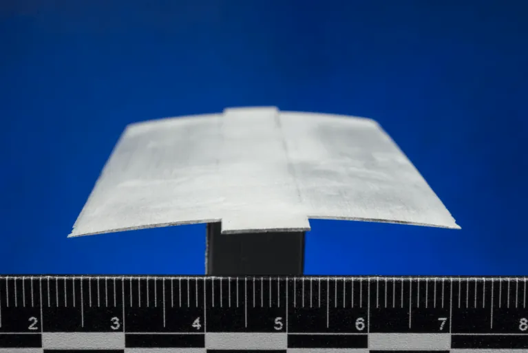 A close-up view of a cross section of a rotor blade, also called an airfoil. The thin, gray material is slightly arched and sits atop a black stand against a ruler.