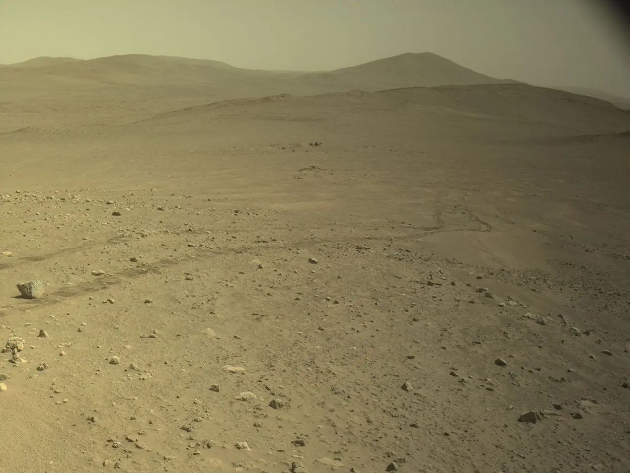A wide view of the Martian surface taken by the Perseverance rover. The foreground features an arid, tan-colored terrain scattered with rocks and pebbles of varying sizes. Faint, parallel rover tracks