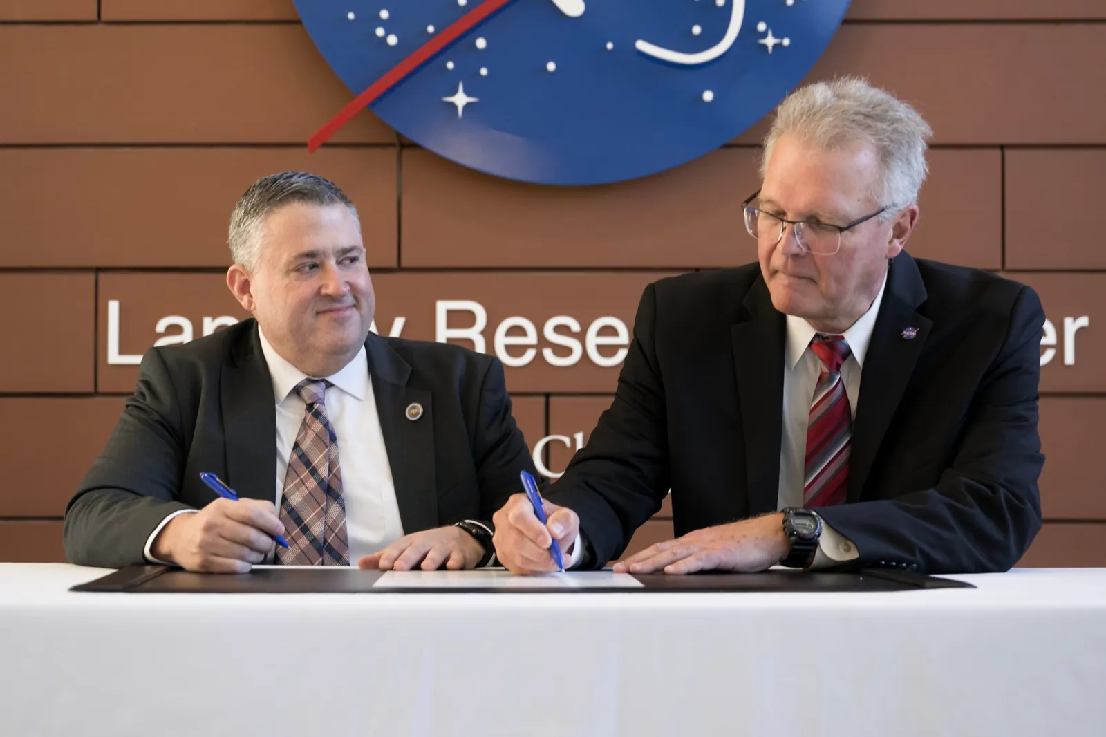 The image shows two men in suits looking down and signing a document on a table. The background wall shows the NASA logo and lettering that spells Langley Research Center.