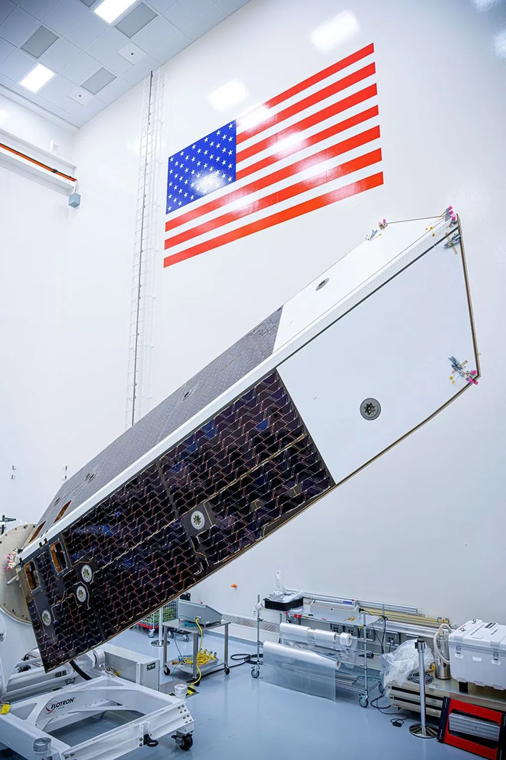 A large spacecraft sunshade and solar array panel rests angled upward on a test rig inside a high bay clean room, positioned beneath a large American flag on the wall.