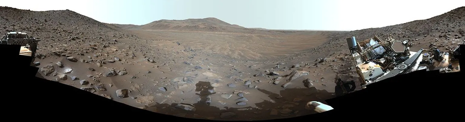 A panoramic view of a barren, rocky landscape with distant hills, framed by parts of a robotic rover visible on the left and right edges.