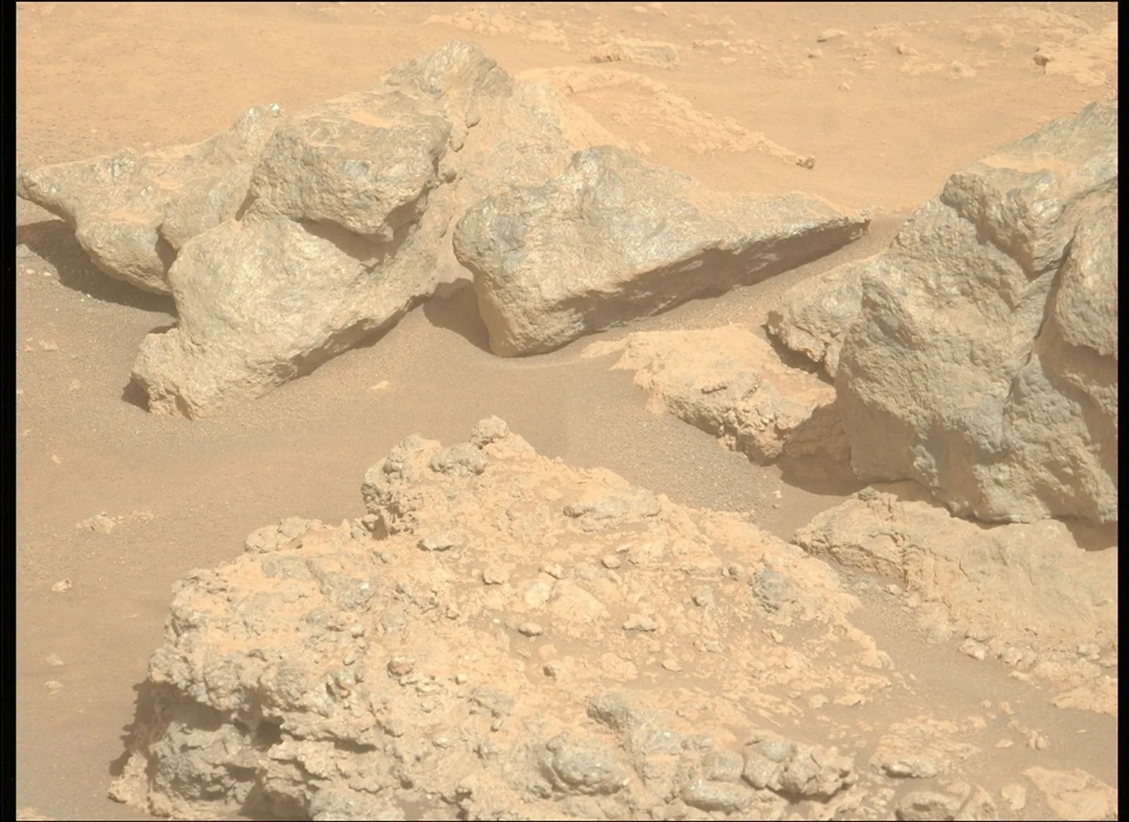 A photograph of the Martian landscape captured by the Perseverance rover. The image shows rough, heavily textured, light-gray and tan rocks of various sizes resting on a dusty, reddish-orange sandy su