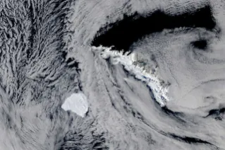 A large, blocky iceberg sits in dark ocean water just southwest of a long, narrow, snow-capped island. Thin clouds partly veil the scene.