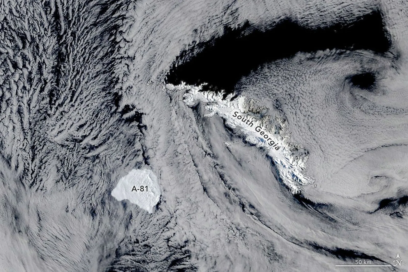 A large, blocky iceberg sits in dark ocean water just southwest of a long, narrow, snow-capped island. Thin clouds partly veil the scene.