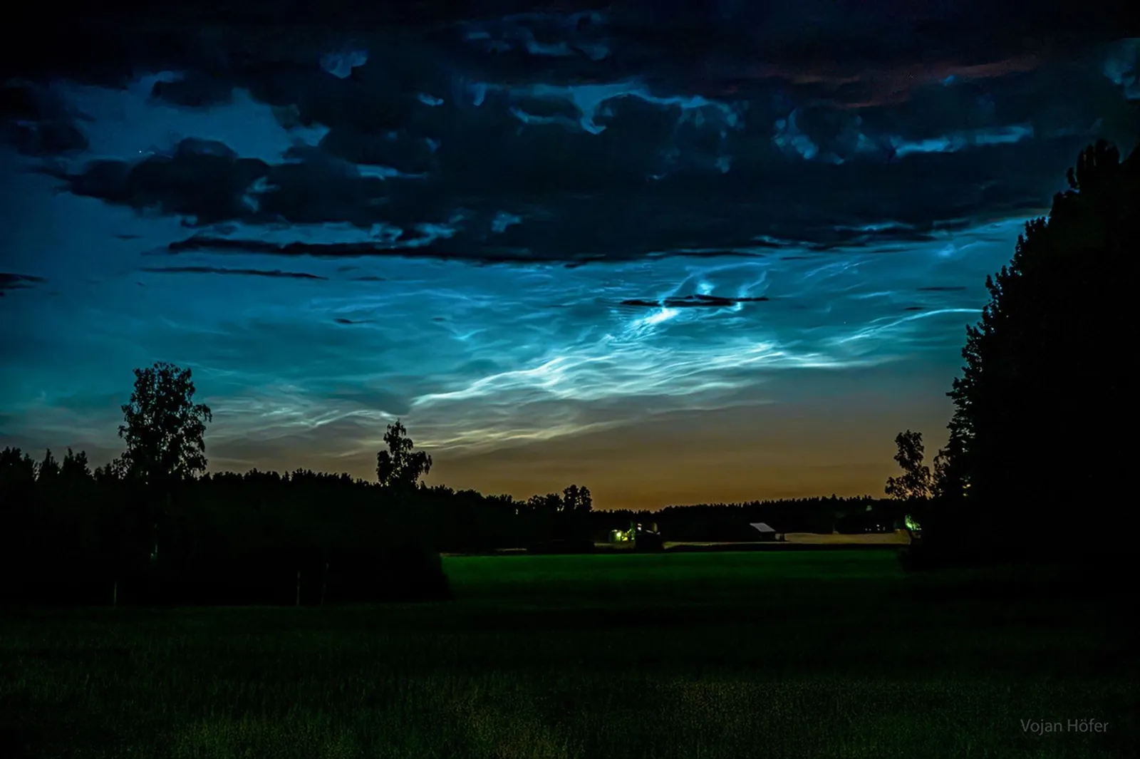 A dark night sky is pictured over a green field bounded by trees in silhouette. Across the image middle are blue filamentary glows -- noctilucent clouds. Dark clouds are seen across the top of the ima