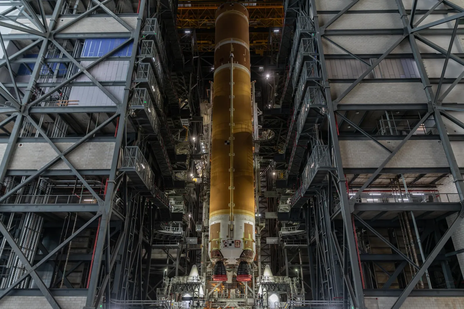 The 212-foot core stage of the Space Launch System rocket is vertical in High Bay 3 of the Vehicle Assembly Building. The nose cones of the two side boosters are visible as the crane places the core s