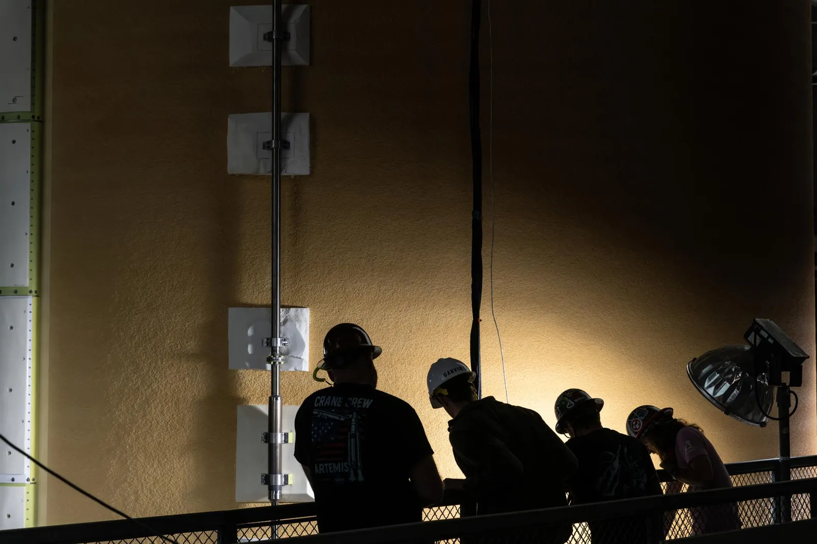 Four people in hard hats are silhouetted against the side of the core stage of the Space Launch System rocket as it is raised in the transfer aisle of the Vehicle Assembly Building. A spotlight on the