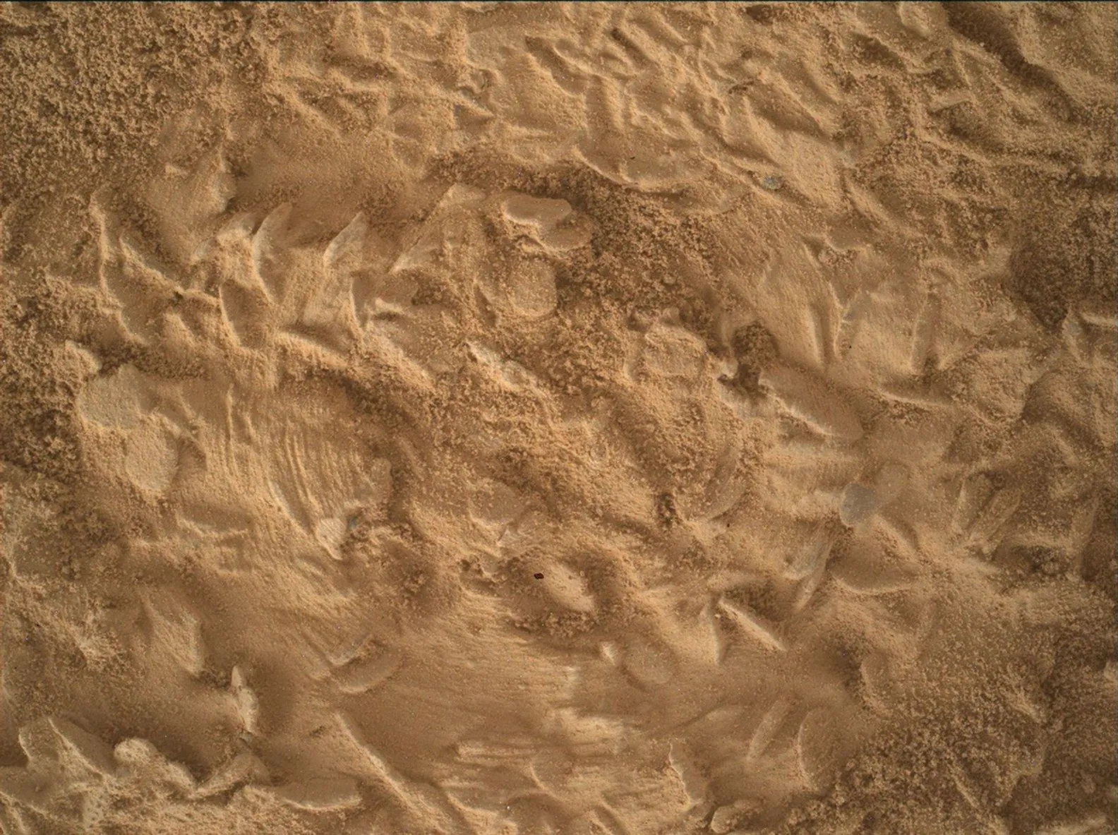 A close-up, overhead view of the Martian surface taken by the Curiosity rover. The image focuses on fine, granular, orange-brown sand that has been heavily disturbed. It features repeating curved ridg
