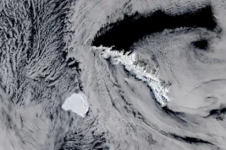 A large, blocky iceberg sits in dark ocean water just southwest of a long, narrow, snow-capped island. Thin clouds partly veil the scene.