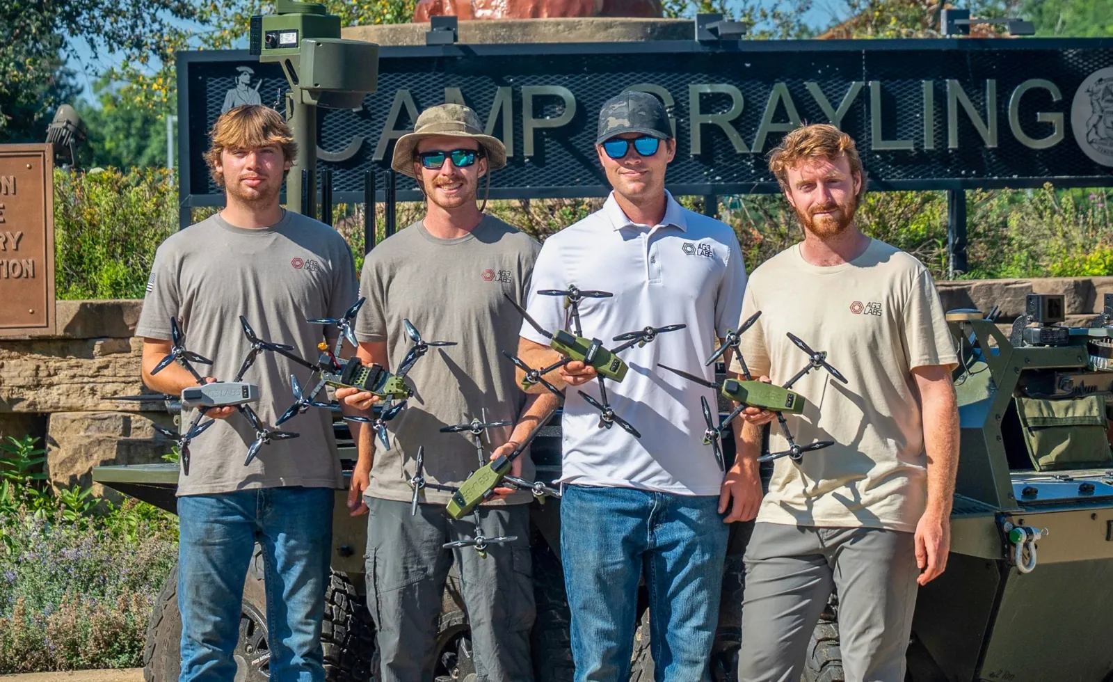 NASA University Challenge image shows four men dressed in t-shirts and jeans, each holding a small quad-copter drone, stand in front of a “Camp Grayling” sign with trees and bushes in the background.