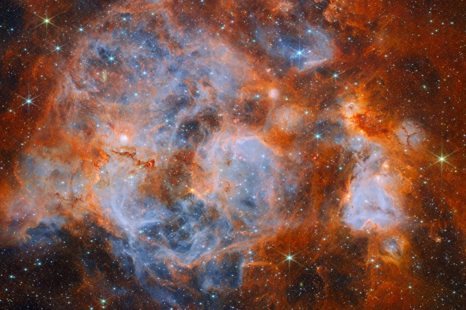 A nebula made of clouds of gas and dust with stars forming within. In the center and out to the left it is filled with thin, wispy blue gas that forms delicate layers. Around this are thicker orange c